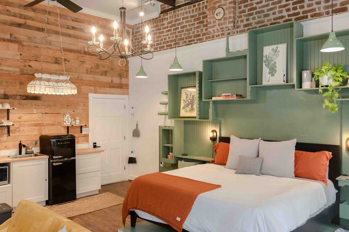 An inviting studio features a spacious bed adorned with a white and orange blanket, complemented by gray and white pillows. Exposed brick walls and wood accents provide warmth, while green shelving adds a modern touch. A kitchenette is visible, enhancing the studio's functionality.