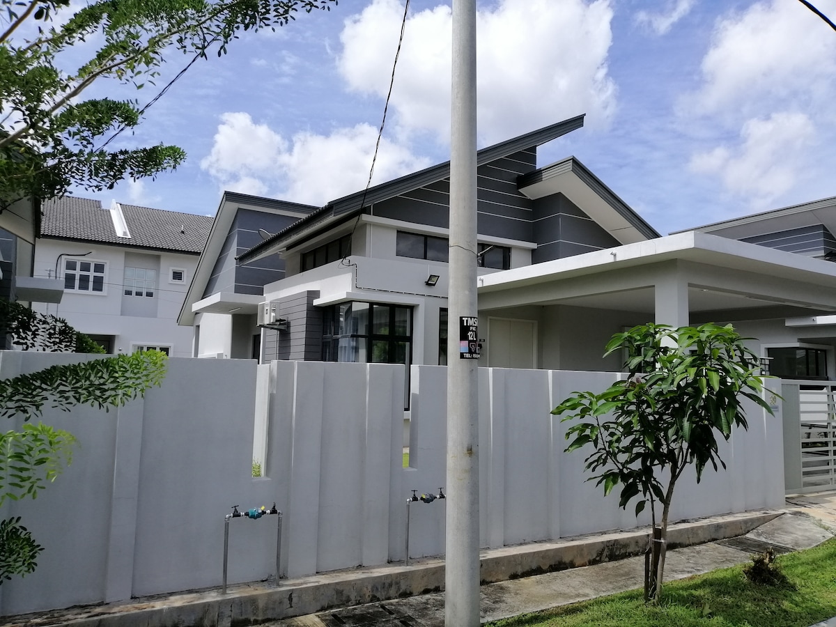 A modern bungalow is depicted, featuring a sleek grey exterior with large windows that provide ample natural light. Surrounded by a well-maintained garden, the property is enclosed by a tall white fence, promoting privacy while maintaining a welcoming presence.
