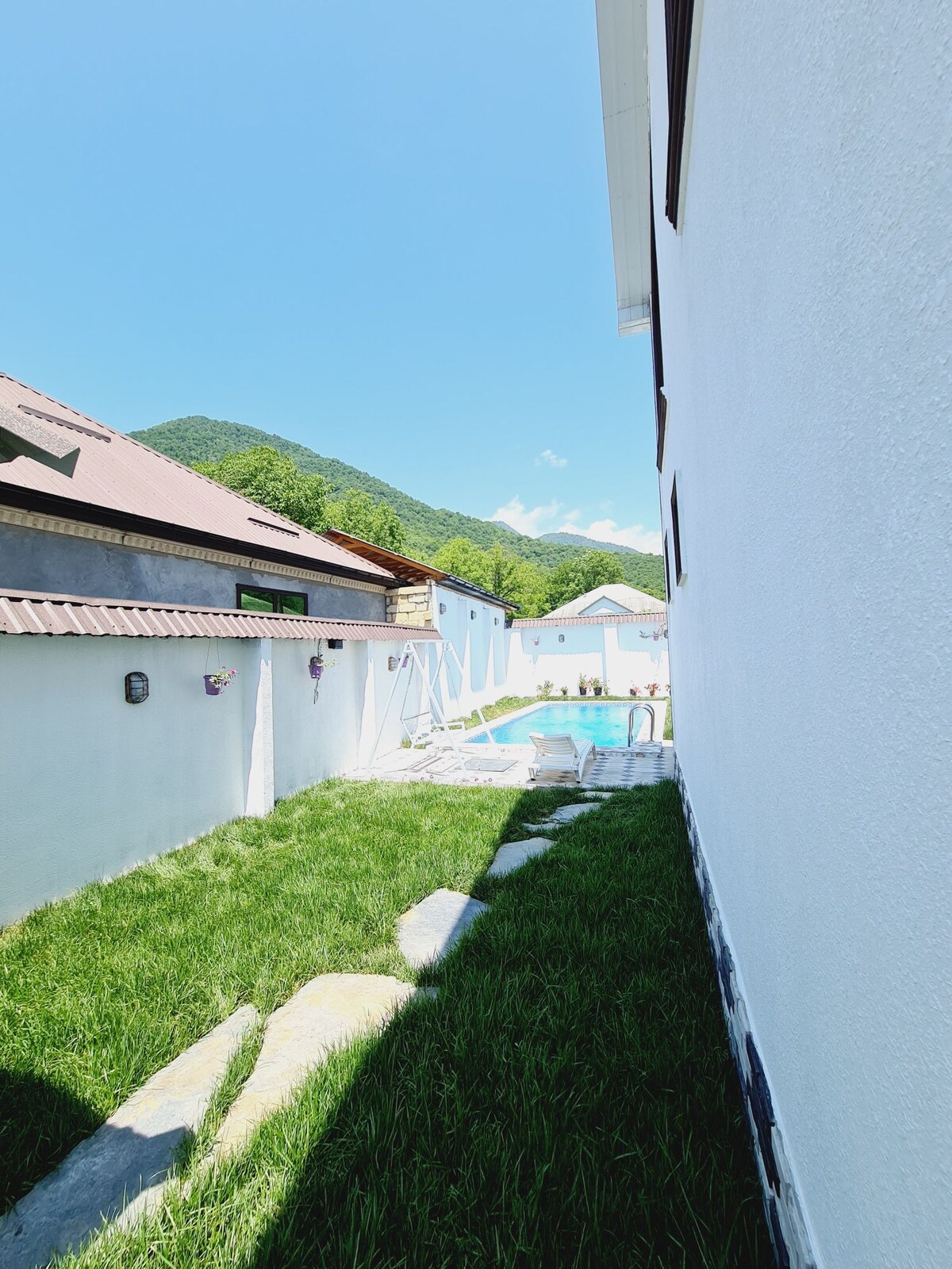A narrow walkway is lined with smooth stones and bordered by lush green grass. The view extends towards a swimming pool, framed by white walls, with mountains visible in the background under a clear blue sky.