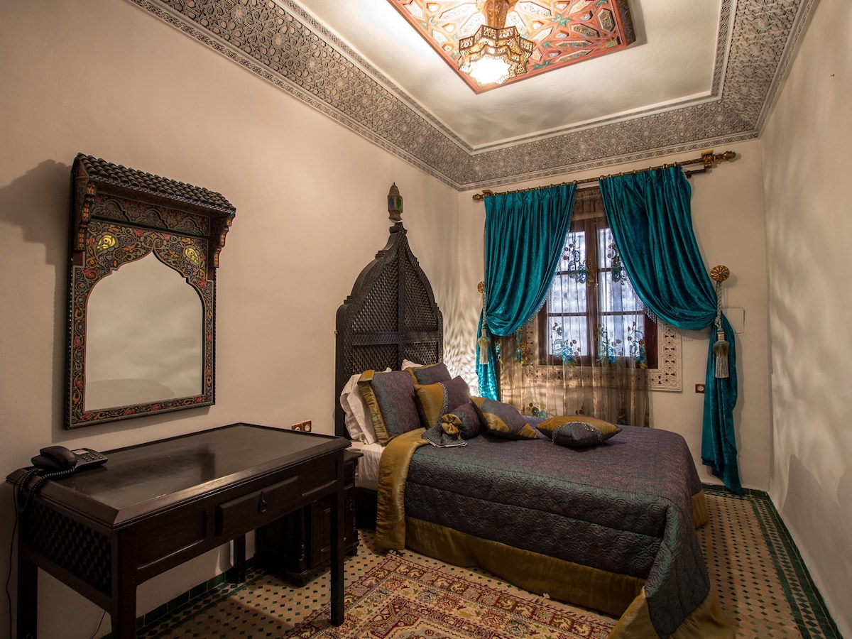 A vibrant bedroom showcases a spacious bed adorned with decorative pillows. Rich turquoise curtains frame a window with intricate patterns, while a handcrafted mirror reflects traditional Moroccan aesthetics. The ceiling features detailed hand-painted designs, enhancing the cultural ambiance of the room.