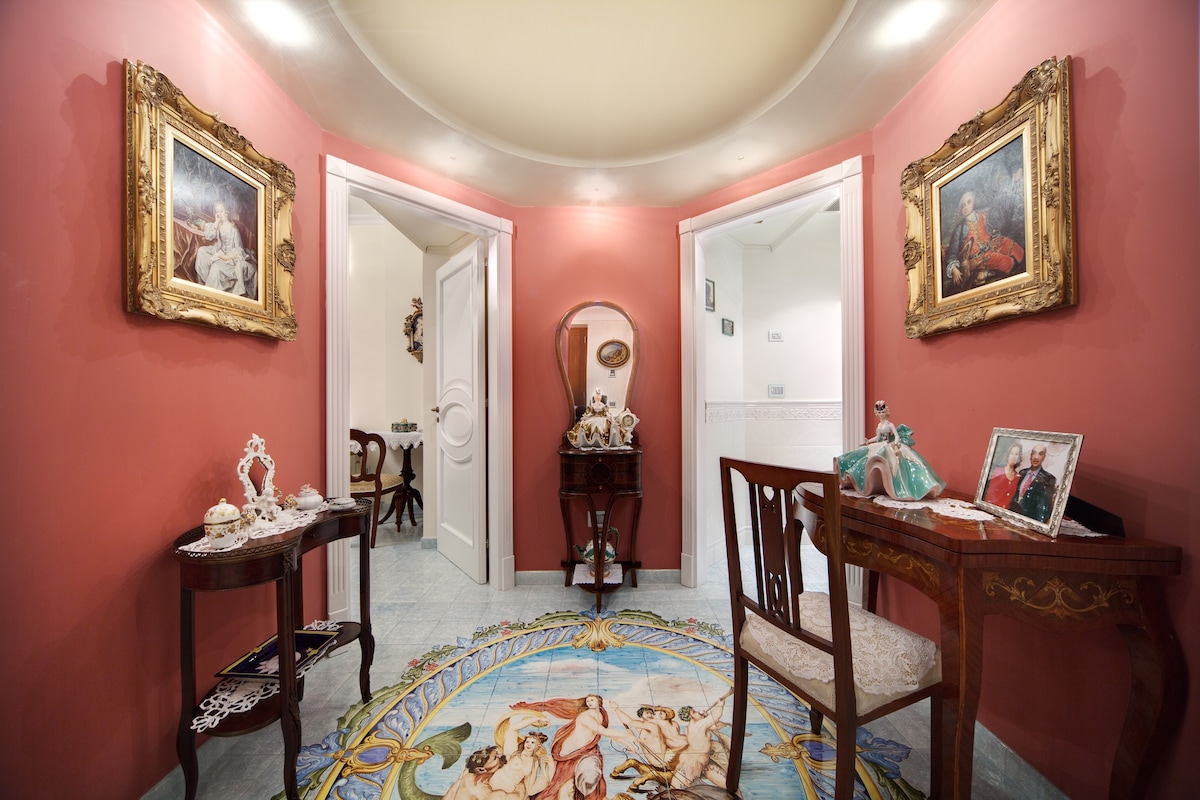 A welcoming entrance hall features a circular ceiling and pink walls adorned with ornate frames containing classical artwork. A round, intricately designed rug lies on the floor, complementing a small wooden table and two elegant chairs. Double doors lead to inviting rooms beyond.