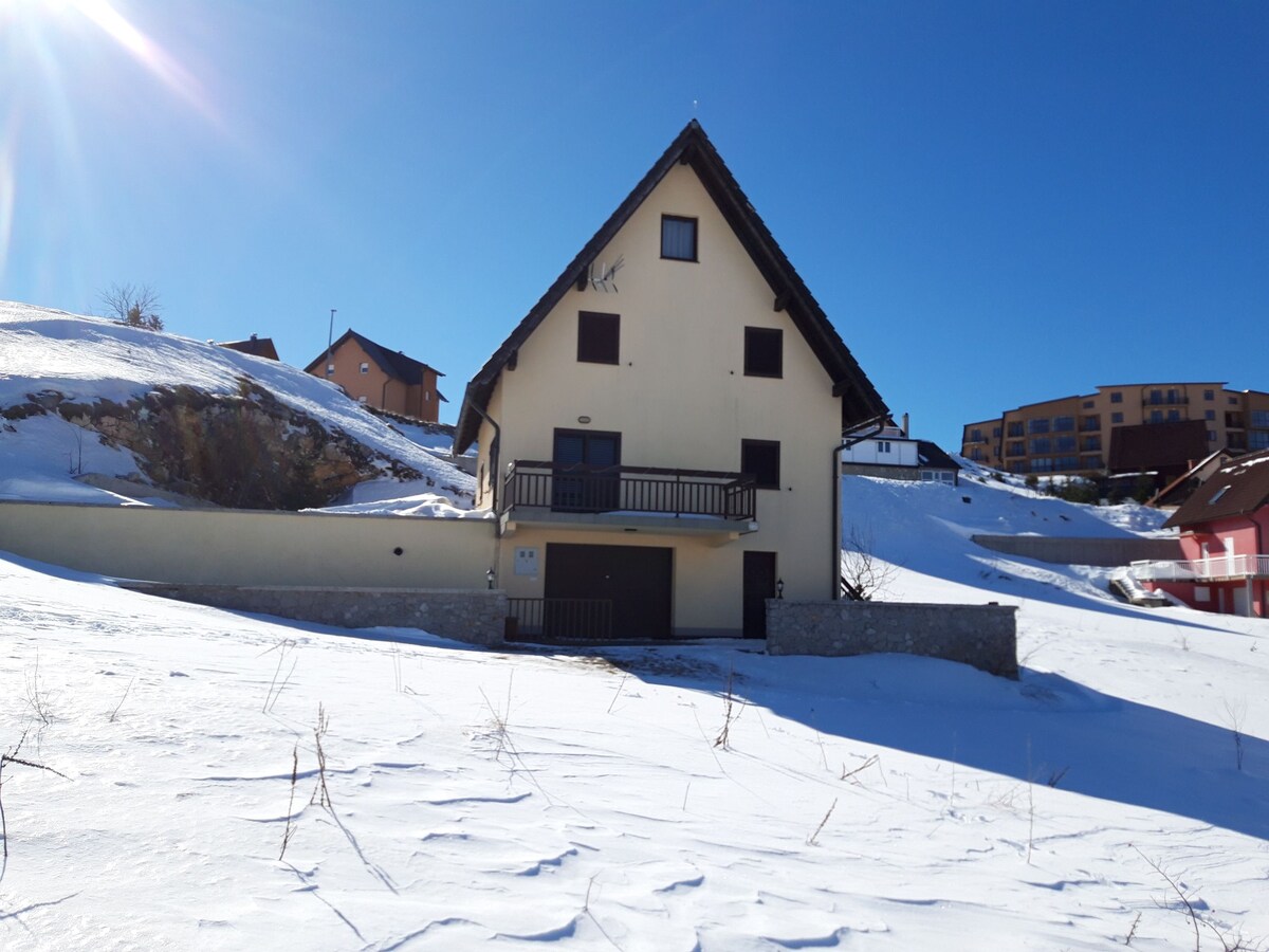 A three-story holiday home stands against a clear blue sky, surrounded by a snow-covered landscape. The building features a steeply pitched roof, multiple balconies, and a stone foundation. Nearby structures are visible, enhancing the picturesque winter scene.