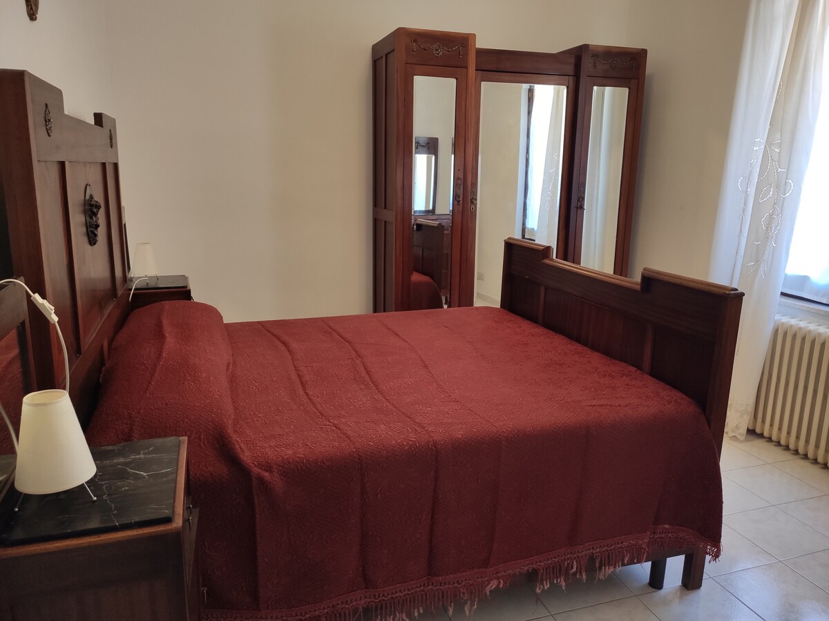 A spacious bedroom is presented with an elegant wooden headboard and a deep red bedspread. A mirror wardrobe with glass doors reflects natural light, while a small bedside lamp adds a subtle touch. Light curtains frame a window, enhancing a cozy ambiance.