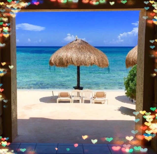 A spacious entryway frames a scenic view of a sandy beach. A thatched umbrella provides shade over two lounge chairs, set against the vibrant blue waters of the Caribbean Sea. Bright skies host a few scattered clouds, enhancing the tranquil setting.