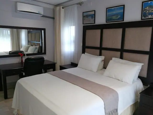 A cozy sleeping area is featured with a spacious bed adorned with a light duvet and neutral-colored throw. A wooden desk and chair are positioned nearby, and framed artwork hangs on the walls. Natural light is softly filtered through sheer curtains.