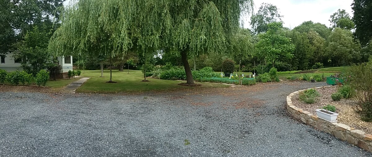 A panoramic view captures the spacious outdoor area, featuring several lush green trees and a well-maintained garden. Gravel pathways meander through the landscape, leading to various shrubs and flowerbeds. The serene setting offers a sense of calm and tranquility, ideal for relaxation.