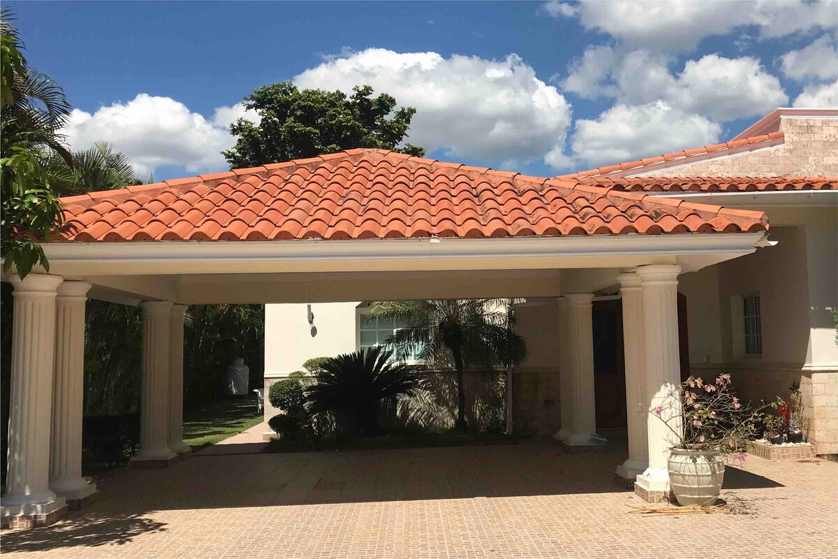 The entrance features a covered carport supported by tall, white columns. A textured tile floor extends throughout the area, with lush greenery and palm plants visible in the background. The roof is adorned with terracotta tiles, creating a warm contrast against the blue sky.