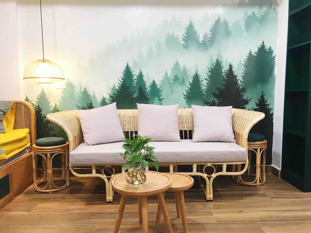 A stylish seating area features a light-colored, woven sofa adorned with soft, neutral cushions. A small round table sits in front, accompanied by decorative greenery. A wall mural showcases a serene forest scene, enhancing the calm atmosphere of the space.