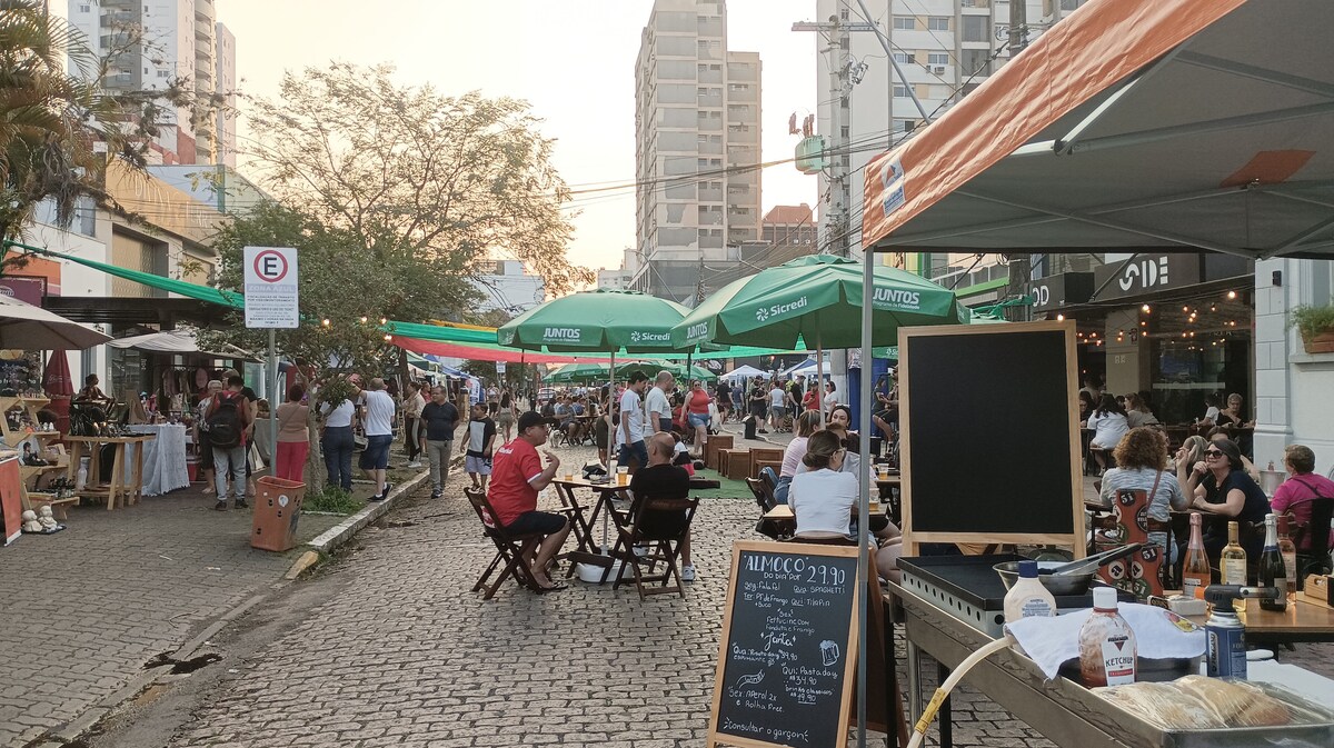 A lively street scene is captured, showcasing outdoor dining areas with green umbrellas providing shade. Rows of tables and chairs are filled with guests enjoying the atmosphere. Buildings rise in the background, indicating a vibrant urban setting.