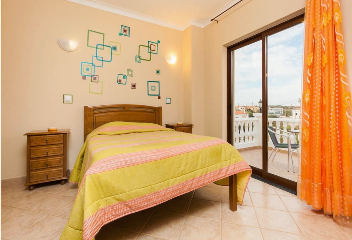 A bright bedroom features a wooden double bed dressed in a colorful striped blanket. Decorative wall frames of varying sizes enhance the space, while large windows allow natural light to fill the room. Outside, a small balcony with chairs is visible.