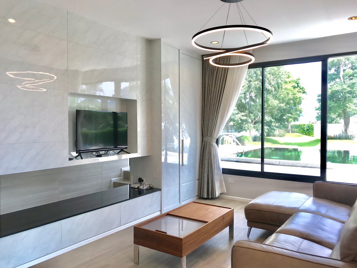 A modern living area features a sleek wall-mounted television and a wooden coffee table. Large windows allow natural light to fill the space, with views of greenery and the swimming pool outside. Soft furnishings create a comfortable setting, with curtains elegantly draping the windows.