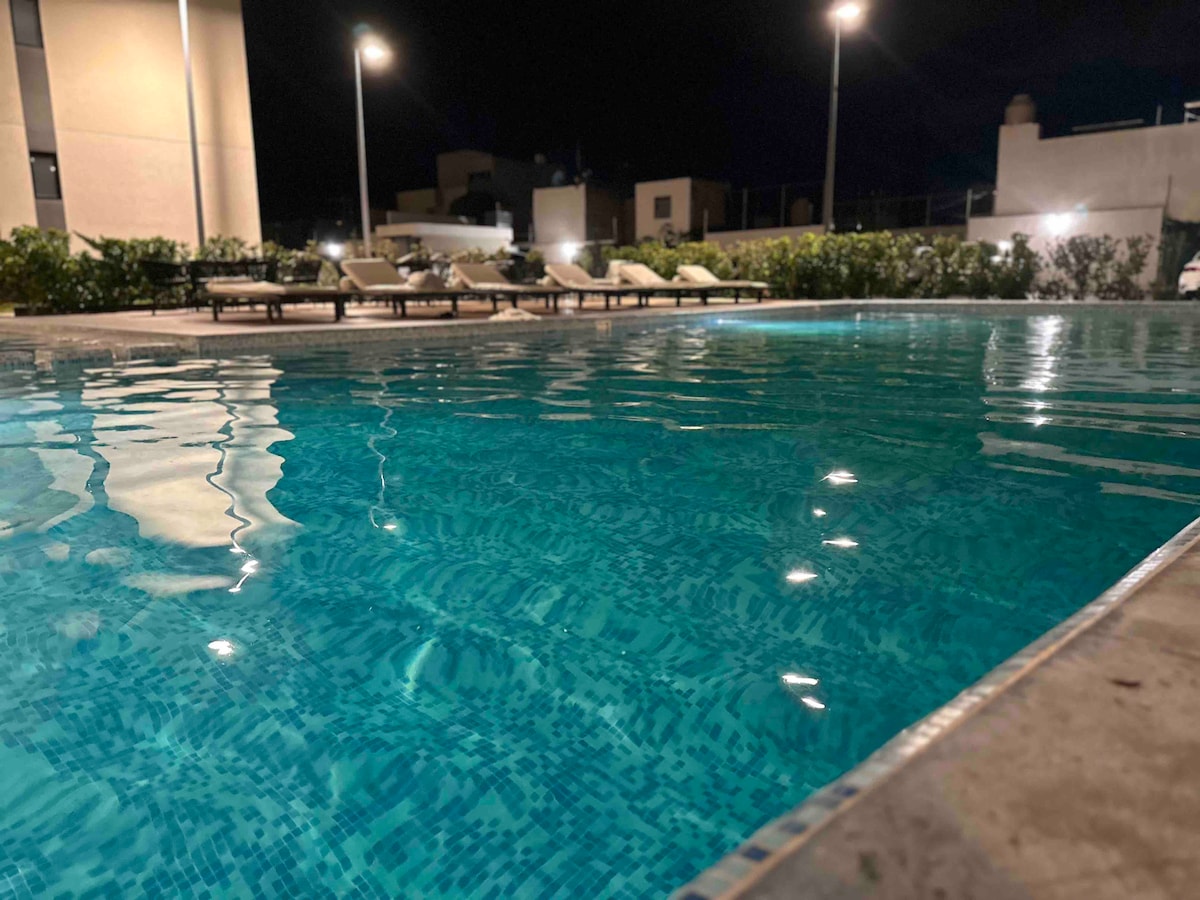 The image depicts a well-lit swimming pool at night, featuring crystal clear water reflecting warm light from nearby lamps. Lounge chairs are positioned along the pool's edge, surrounded by lush greenery that enhances the serene atmosphere.