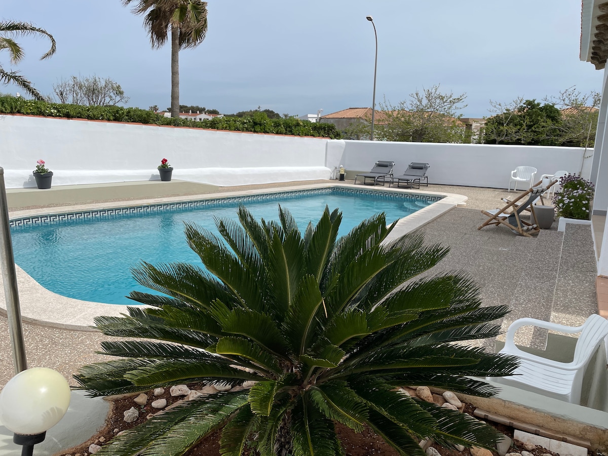 A private swimming pool is framed by a well-maintained garden featuring a palm and potted plants. Sun loungers are positioned beside the pool, while a comfortable chair is seen near the edge. The serene outdoor space is enhanced by a clear sky.