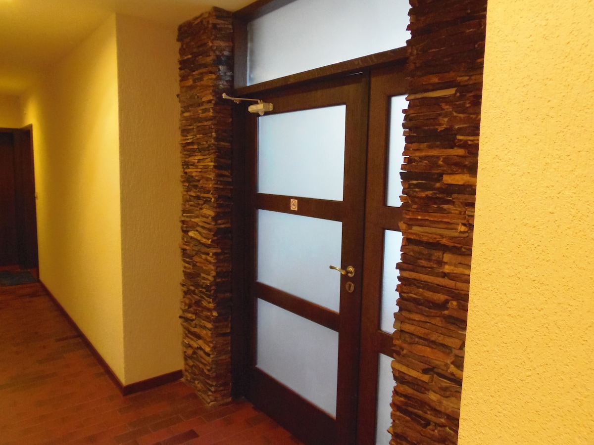 A wooden door with frosted glass panels is framed by textured stone walls, creating a modern yet rustic entrance. The surrounding corridor is softly lit, featuring smooth walls in neutral tones, enhancing the inviting ambiance of the space.