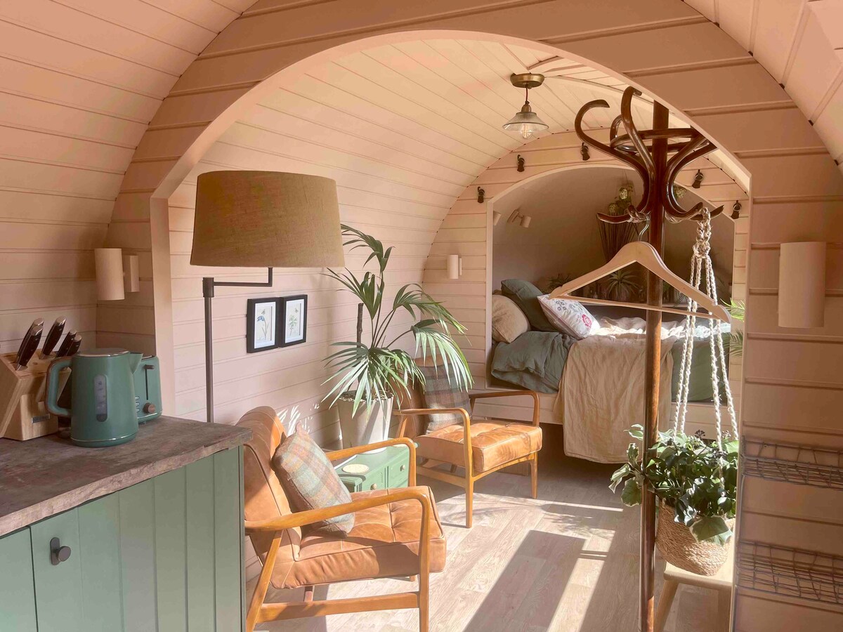 A charming interior space showcases an arched ceiling with soft pastel tones. Seating includes two wooden chairs and a hanging botanical arrangement, while a cozy bed is located in the back section. Natural light filters through the window, illuminating the inviting atmosphere.