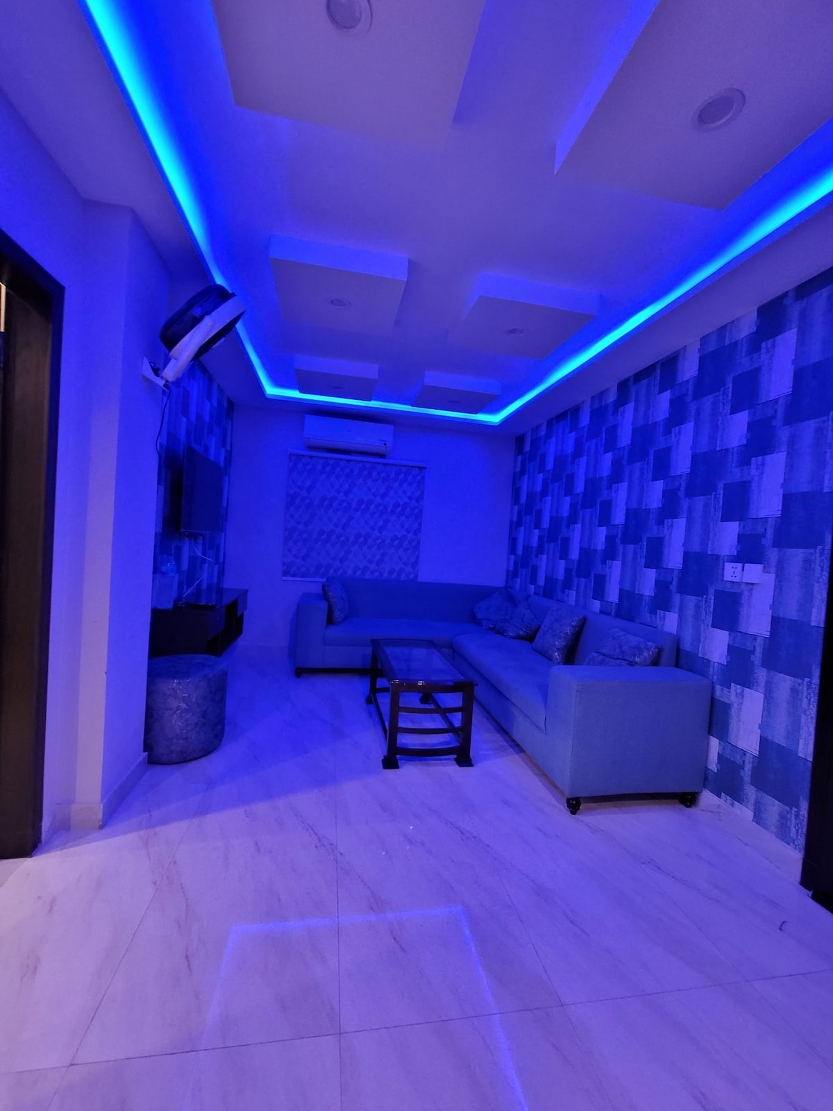 A modern living area is illuminated by blue LED lighting along the ceiling, highlighting textured blue walls. A light blue sectional sofa is positioned against one wall, accompanied by a small dark coffee table in front. A window shade partially filters natural light.