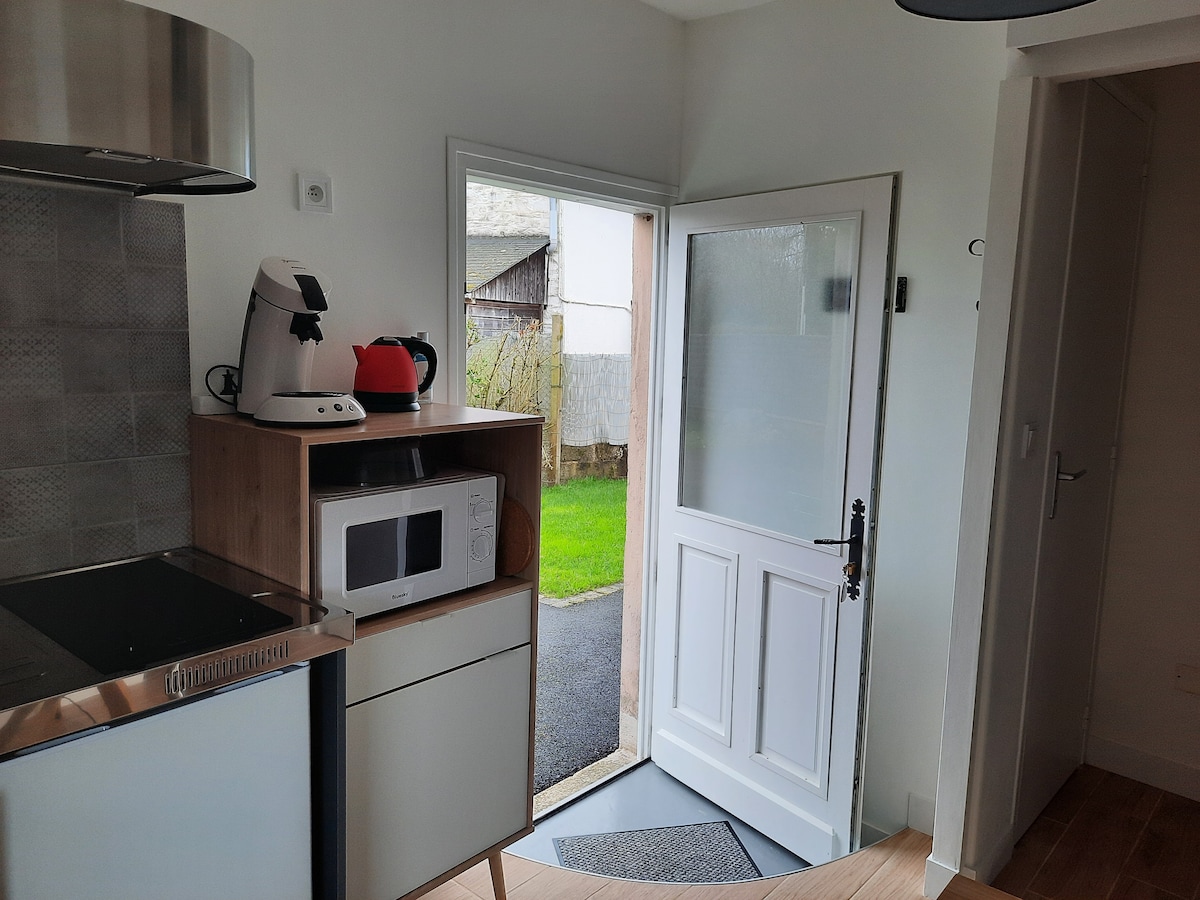 A view from the kitchen area towards the entrance, featuring a door opened to a garden space. Appliances like a kettle, coffee maker, and microwave are visible on the countertop, with a smooth transition to the outdoor area showcased through the door.