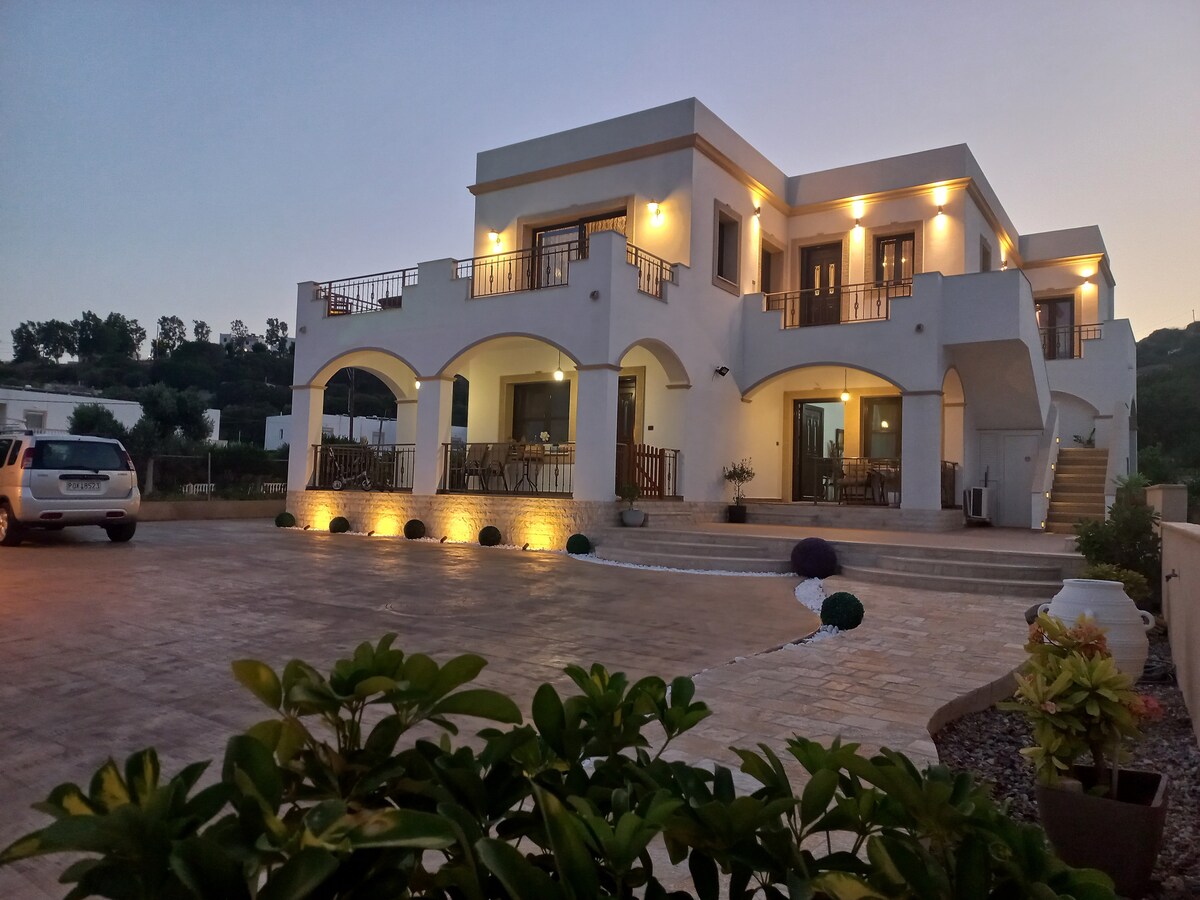 A two-story house features spacious balconies and arched doorways, elegantly illuminated in the evening. The surrounding patio is adorned with potted plants, and ample parking is visible in front. The setting highlights the welcoming architecture against a twilight backdrop.