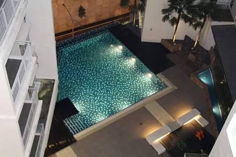 A large swimming pool is surrounded by stone and tiled surfaces. The water reflects light, creating a shimmering effect. Comfortable seating areas with lounge chairs are positioned nearby, accentuated by tropical plants that enhance the outdoor atmosphere. A smaller pool is visible to one side.