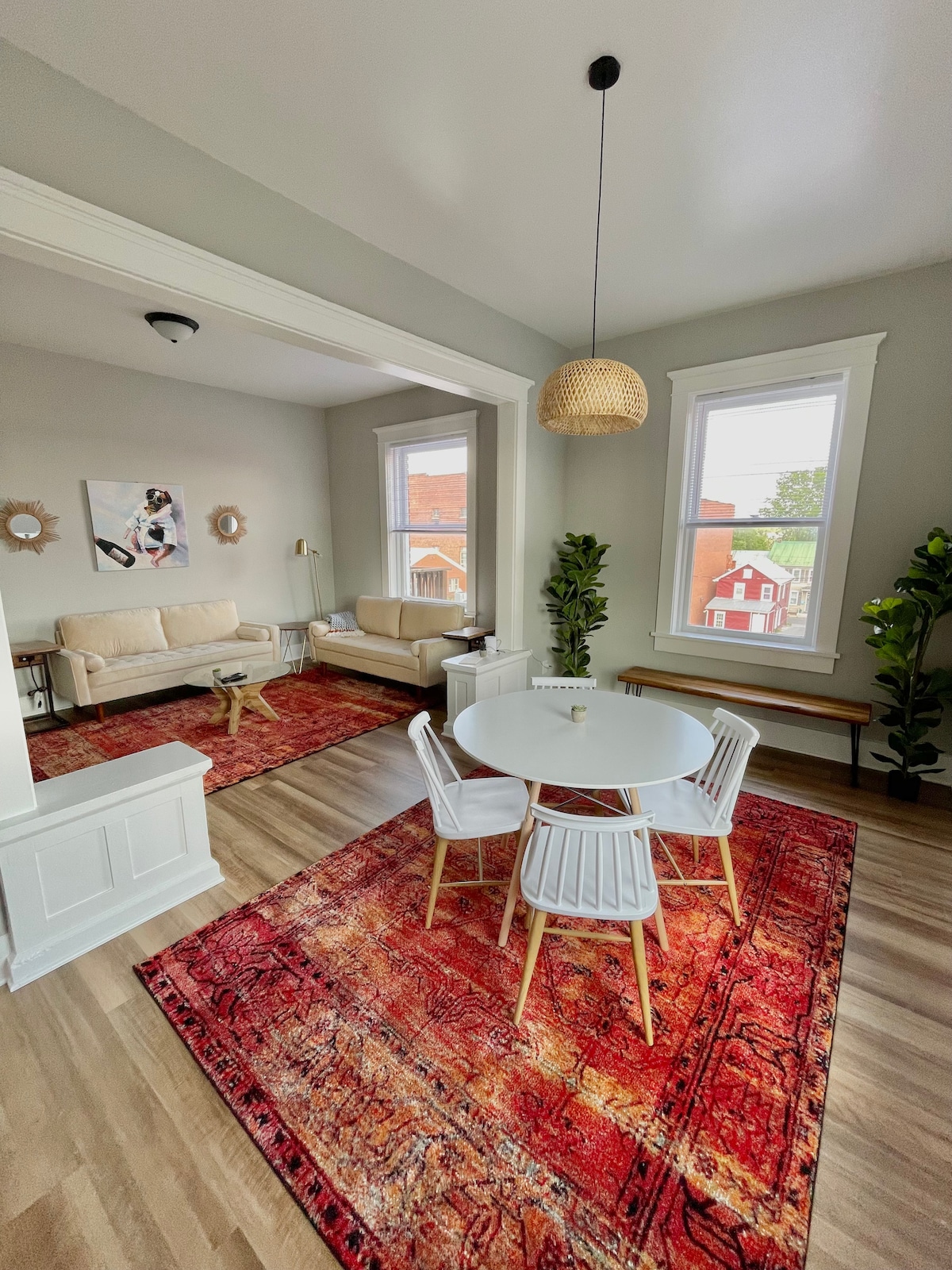 The living area features a dining table surrounded by four white chairs, situated on a vibrant red area rug. Two cream-colored couches are positioned against the walls, with large windows letting in natural light. Decorative elements include a pendant light and potted greenery.
