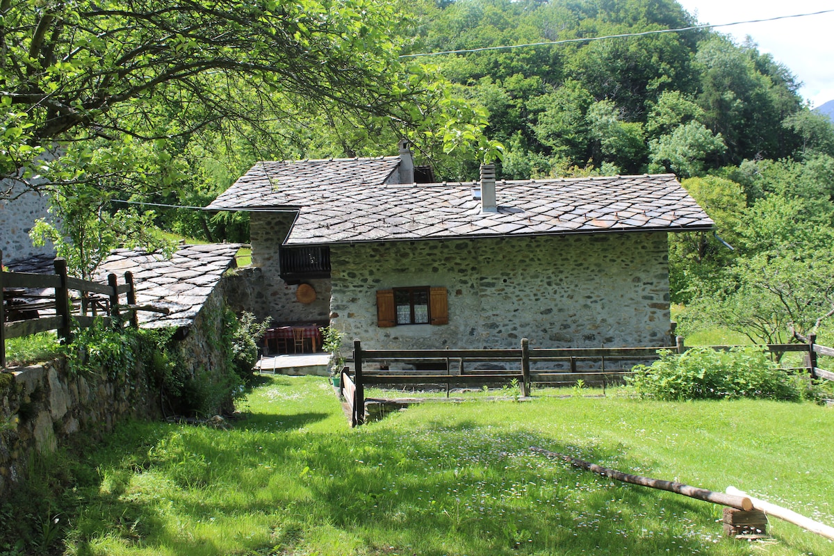 A rustic stone building with a traditional slate roof is set against a backdrop of lush greenery. Features include wooden shutters on the windows and a small outdoor seating area. A grassy expanse surrounds the structure, enhancing its natural setting.