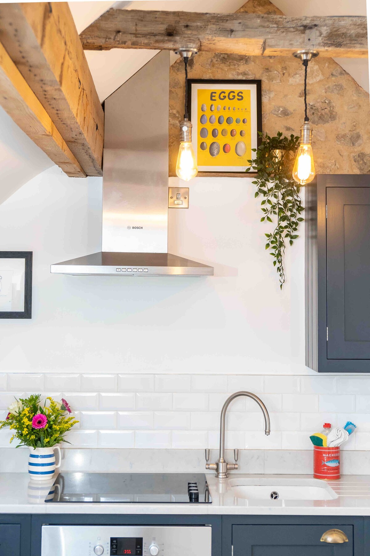 The kitchen area features wooden beams across the ceiling and modern appliances, including a stainless steel hood above the stove. A small vase of flowers adds a splash of color to the countertop, accompanied by practical cleaning supplies, creating a functional and well-designed cooking space.