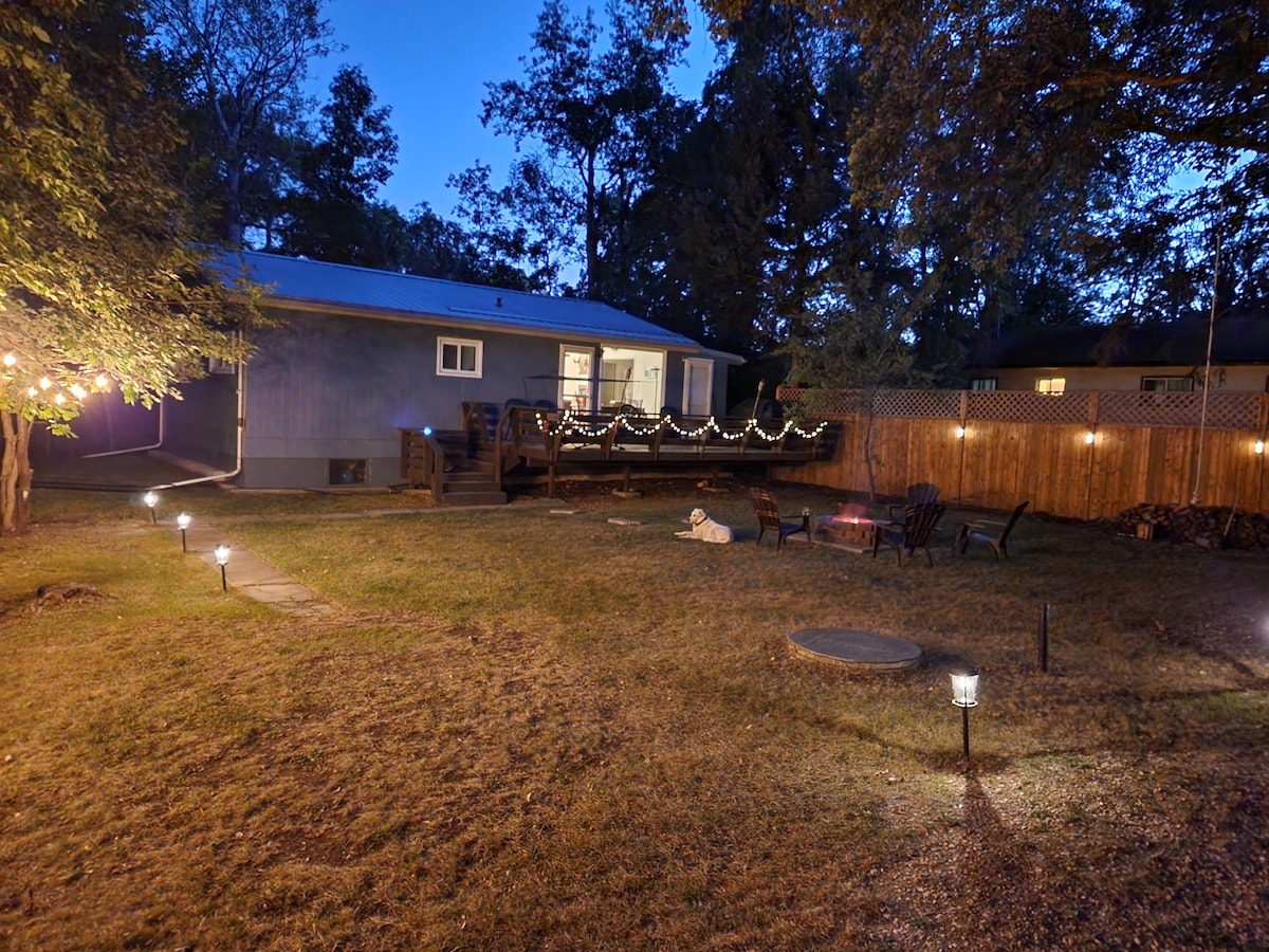 The exterior of the cottage is illuminated at night, with warm lights adorning the deck and surrounding trees. The landscaped yard features a fire pit area, with outdoor seating positioned around it. Pathway lights guide the way through the grassy space, creating a cozy ambiance.