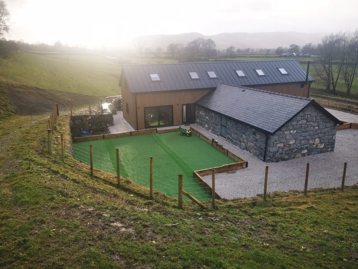 An aerial view captures the contemporary family home, surrounded by lush green fields. The exterior combines stone and modern materials, with large windows reflecting natural light. A manicured artificial grass area provides space for outdoor activities, enclosed by wooden fencing that enhances the privacy.
