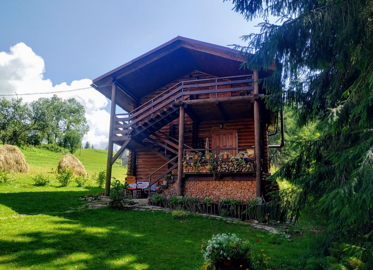 A wooden cabin with a spacious porch stands amidst lush greenery. A staircase leads up to the entrance, surrounded by flower beds and decorative elements. Hay bales are visible in the background, emphasizing the rural setting under a partly cloudy sky.