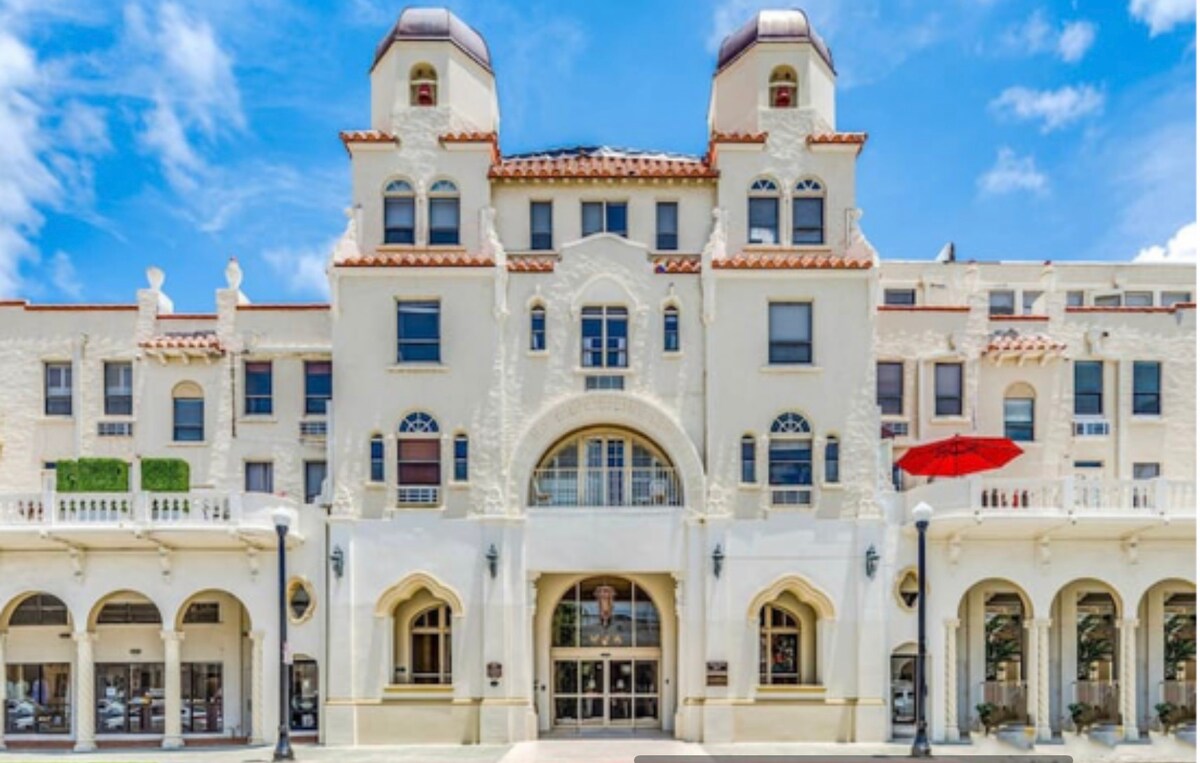 The entrance of the historic Palm Beach Hotel Condominium is showcased, featuring classic Mediterranean architecture. Distinctive towers are topped with rounded domes, while arched windows and decorative details underline the building's elegance. A vibrant red umbrella adds a pop of color to the outdoor seating area.