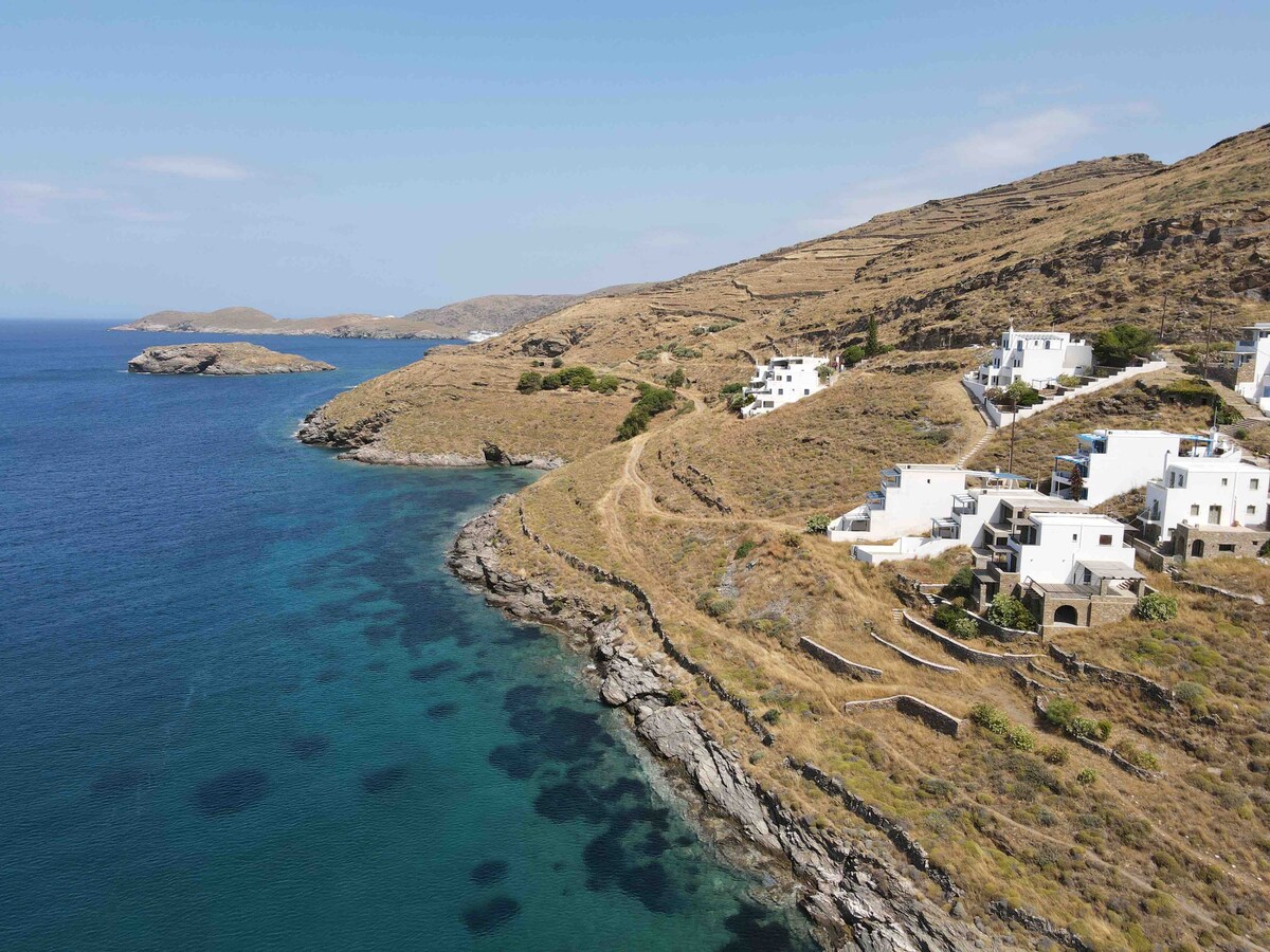 A coastal view reveals a rugged shoreline lined with dry grasses and small white houses with colorful accents. The crystal-clear blue water contrasts with the rocky landscape, while the distant hills complete the serene seascape.