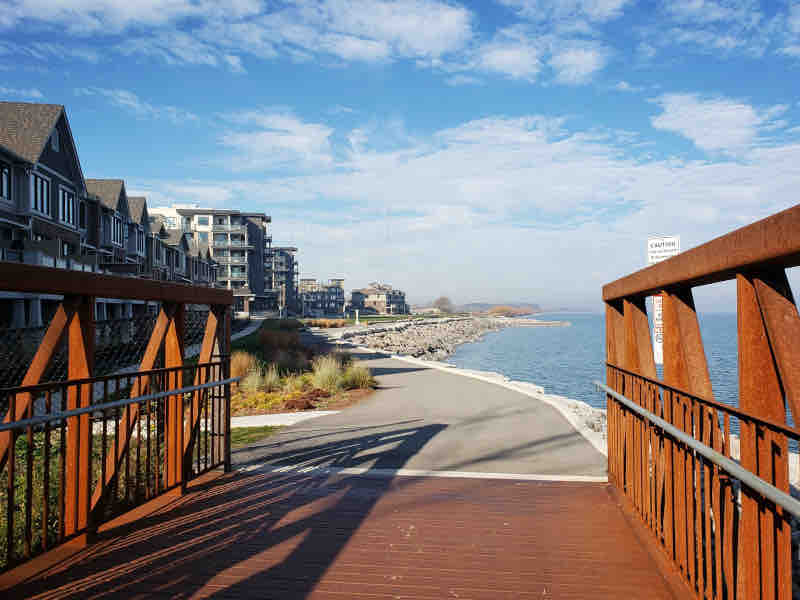 A scenic waterfront view is presented from a wooden bridge, leading to a paved pathway along the shore. Surrounding buildings offer a mix of modern architecture, and the calm water extends into the distance under a clear blue sky. Natural vegetation lines the path.