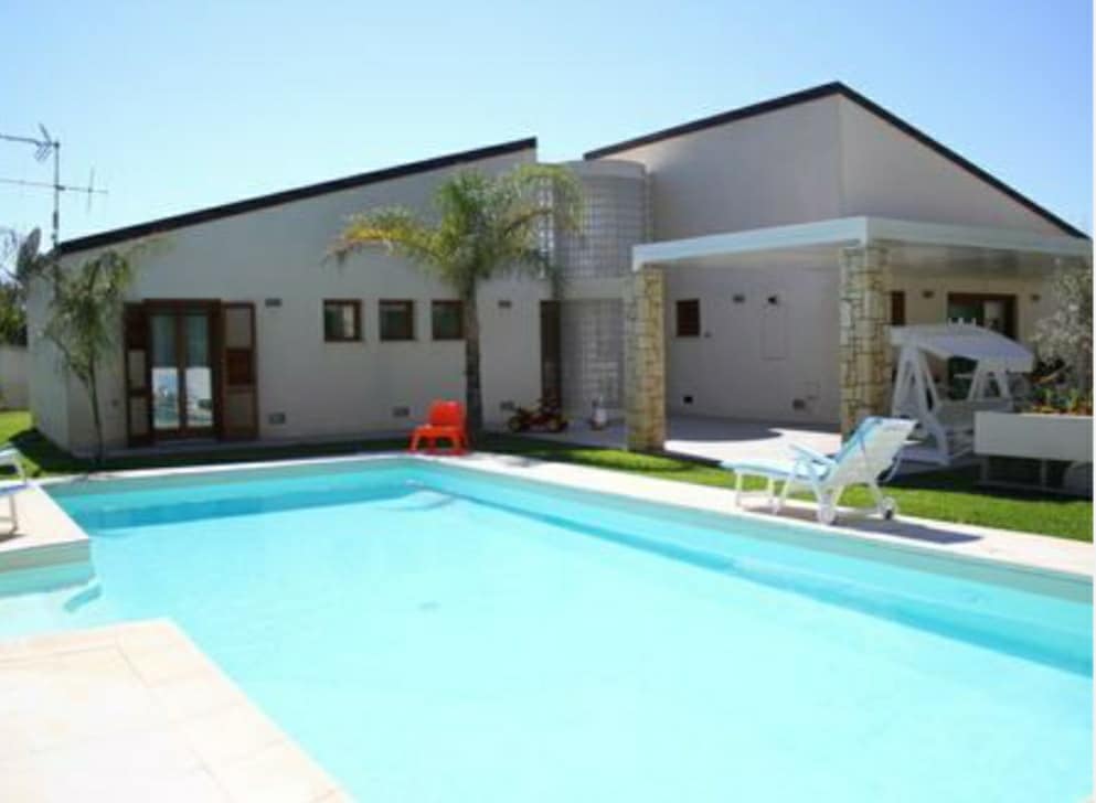 A modern villa is showcased with a private swimming pool surrounded by manicured grass. The structure features large windows and a cozy terrace. Sun loungers and a small orange chair are positioned near the pool, offering a sunny spot for relaxation.