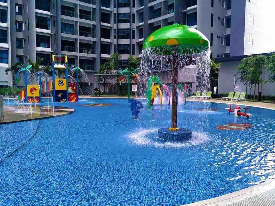 The outdoor swimming pool features a vibrant water playground, complete with a shaded water fountain and colorful play structures. Lush greenery surrounds the area, and lounge chairs are arranged nearby, inviting relaxation. The pool's clear blue water glistens under the bright sunshine.