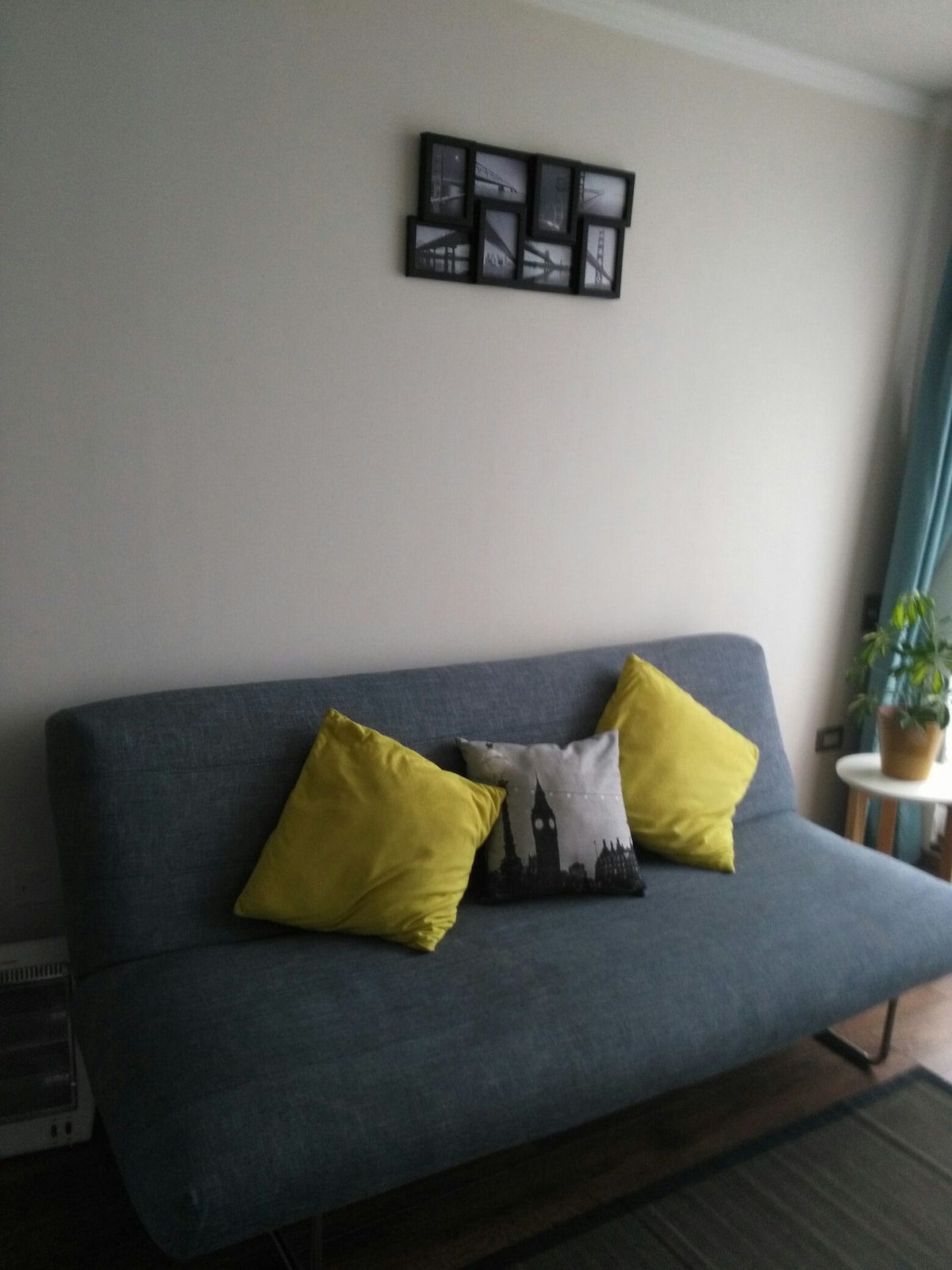 A gray sofa is positioned against a light-colored wall, complemented by two bright yellow cushions. Above the sofa, a framed collage of black-and-white photographs adds character to the space. A small potted plant sits on a light-colored table to the right.