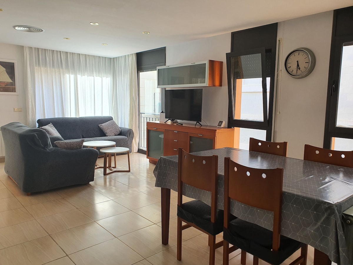 A bright living area features large windows allowing natural light to fill the space. A comfortable sofa is located adjacent to a round coffee table. An entertainment unit with a television stands nearby, while a dining table with wooden chairs is positioned within the open layout.