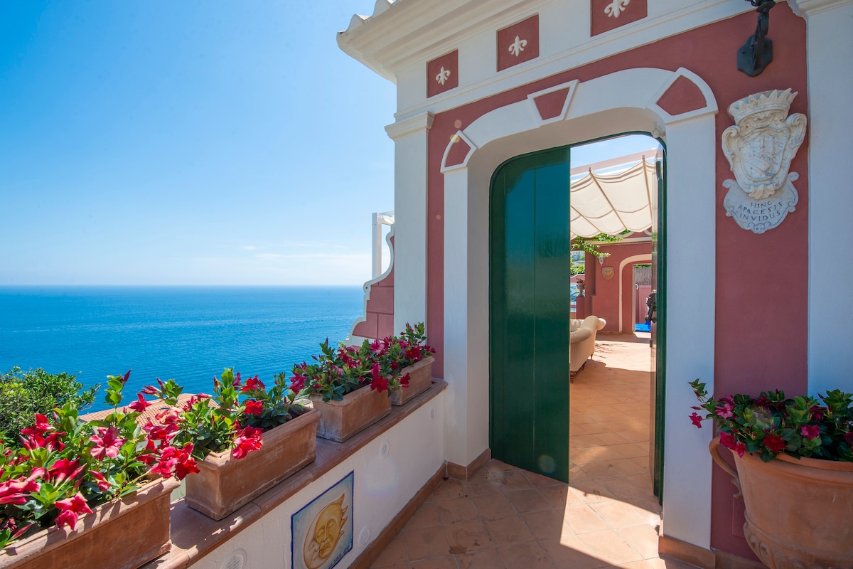 An inviting entrance is framed by vibrant flower pots filled with red blooms. A green door opens to a terrace with expansive views of the blue sea and sky, emphasizing the outdoor connection. The warm tones of the building harmonize with the coastal landscape.