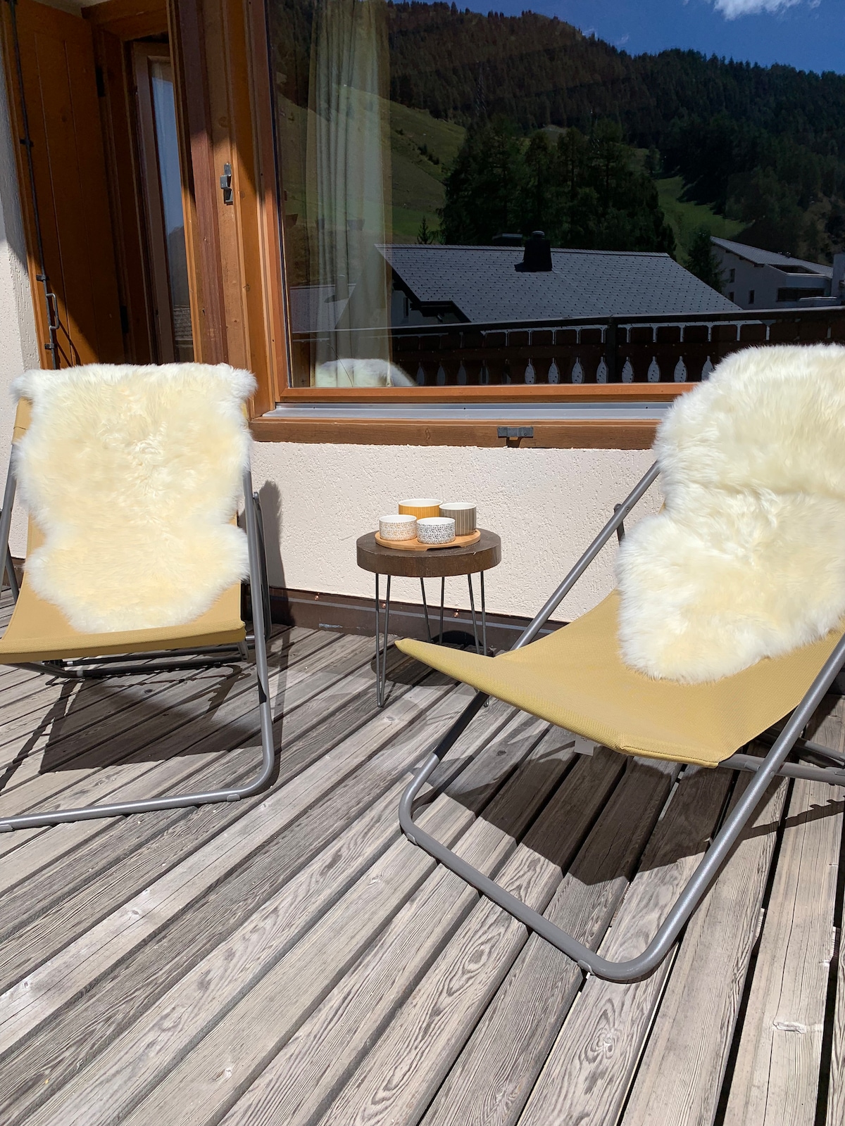 Two loungers with fluffy white cushions are positioned on a wooden terrace. A small round table sits between them, adorned with light-colored dishes. Sunlight filters in through a nearby window, illuminating the space, while the scenic outdoor view provides a relaxing ambiance.