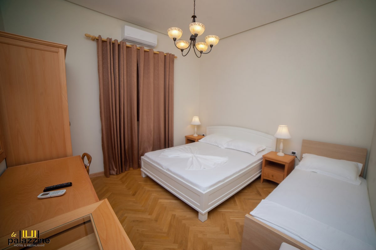 A brightly lit guest room features a spacious double bed and a single bed, both neatly made. Two bedside tables with lamps are positioned beside the double bed. A desk and chair are situated against one wall, and warm curtains partially cover the window.
