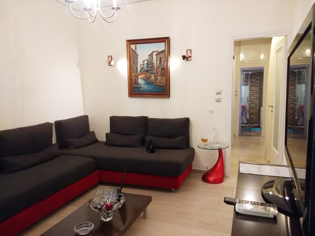A spacious living area features a large corner sofa in dark fabric, complemented by a vivid red accent. A wooden coffee table sits in front, with a decorative glass bowl present. A vibrant painting of a canal scene hangs on the wall, while a flat-screen television is positioned nearby.