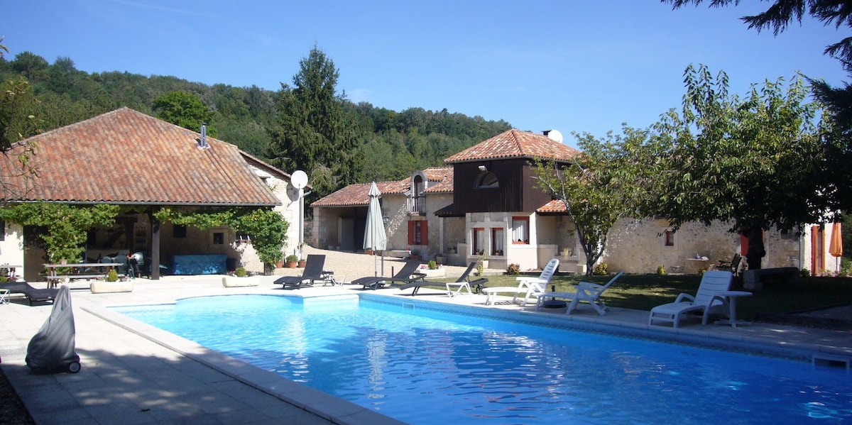 A large swimming pool is surrounded by sun loungers and green grass. Several buildings with terracotta roofs are visible in the background, set against a backdrop of trees. The area appears spacious and inviting, ideal for relaxation.