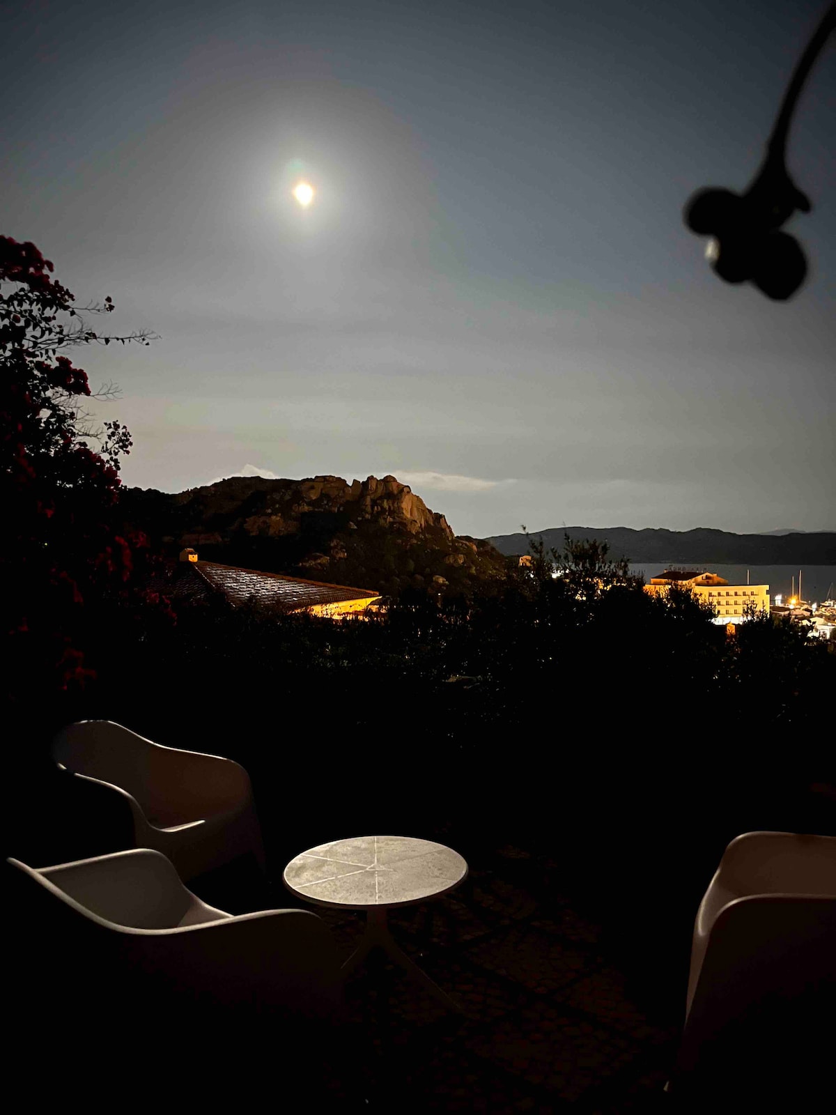 A serene outdoor view is presented, featuring two lightweight chairs and a small round table. The moonlight casts a gentle glow over surrounding foliage and nearby rocky hills. In the background, shimmering water reflects the night sky, creating a tranquil atmosphere.