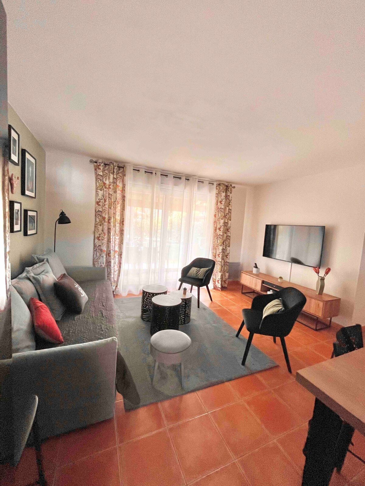 A cozy living area is furnished with a grey sofa and two black chairs. A round coffee table sits at the center, surrounded by natural light flowing through large windows draped with floral curtains. A TV is mounted on the wall across from the seating arrangement.