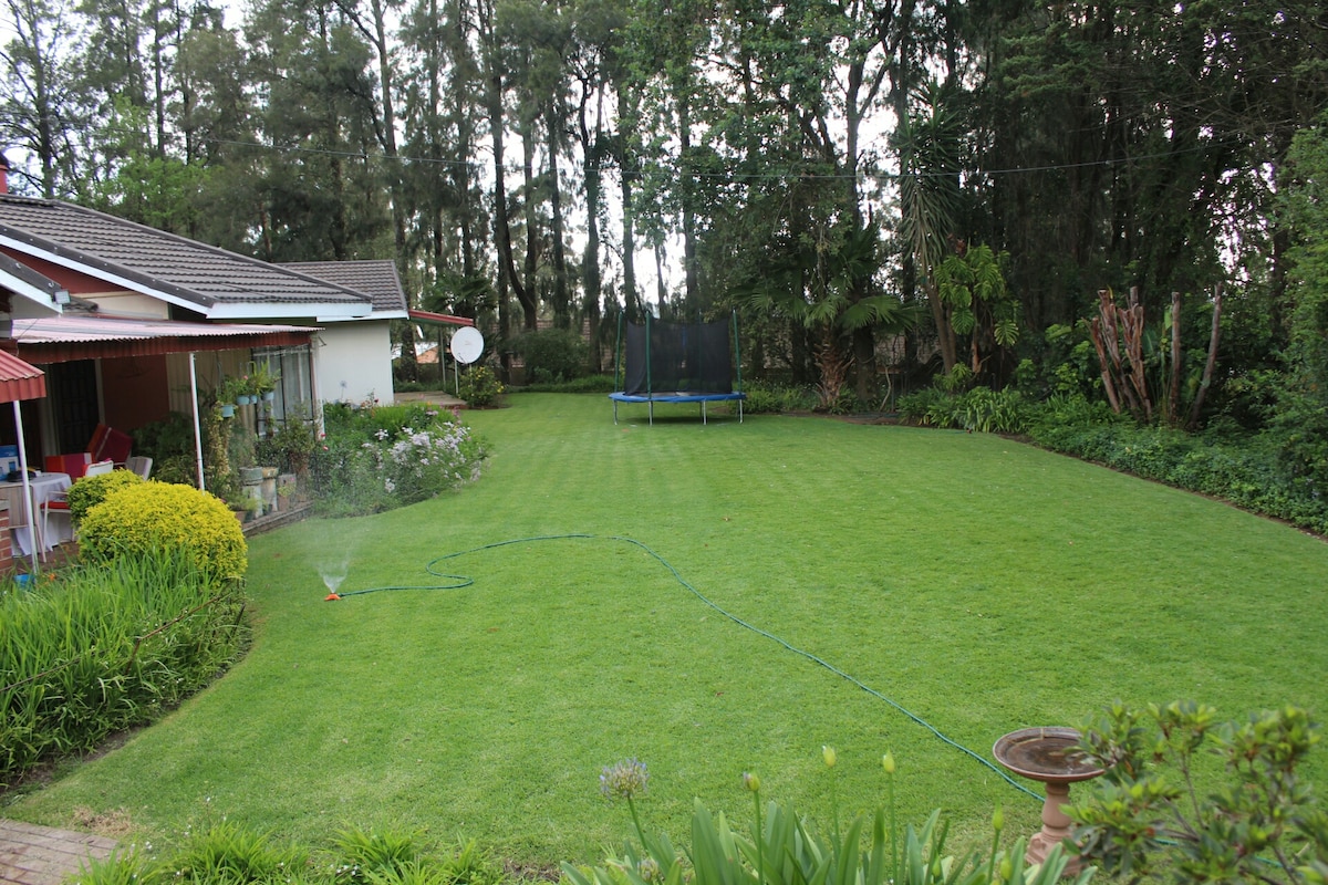 A spacious lawn is surrounded by trees, providing a natural backdrop. A trampoline is visible in the background, along with a small fountain in the foreground. The lawn is well-maintained, and a sprinkler is active, ensuring the grass remains lush.