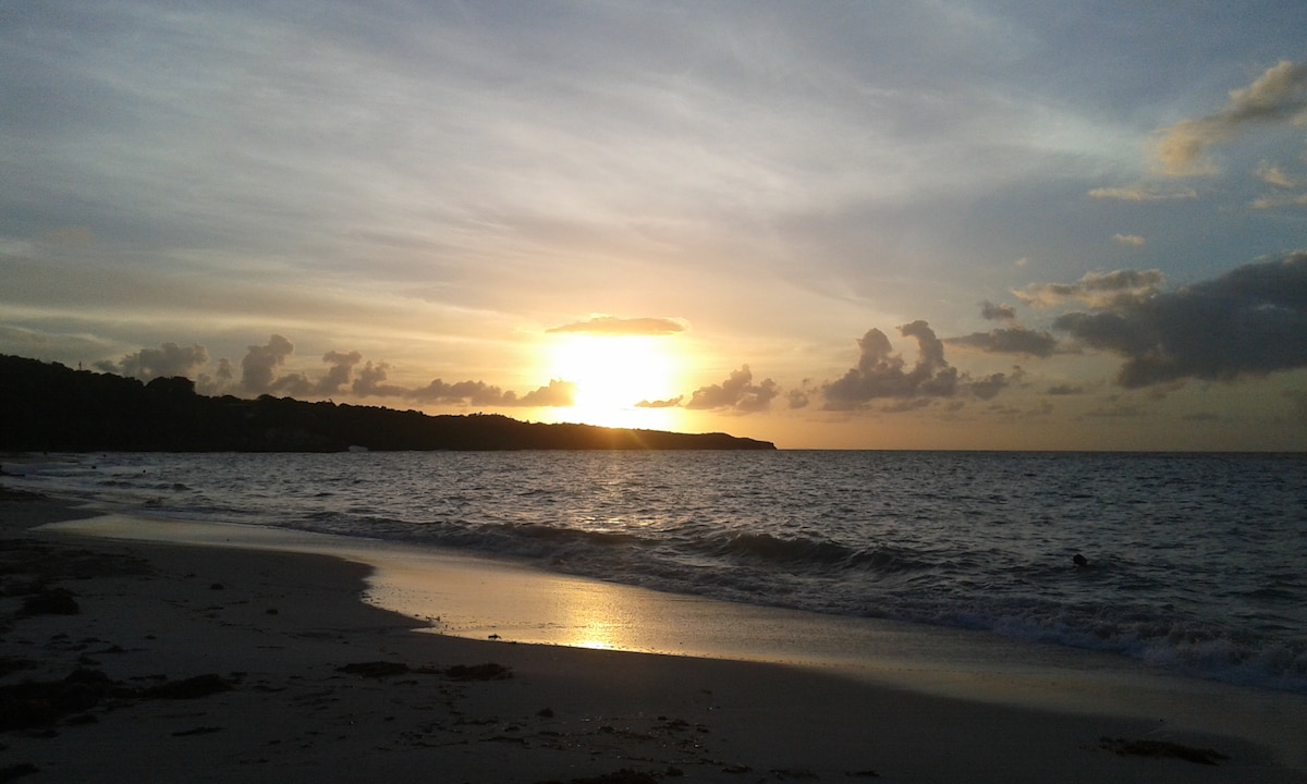 A serene beach scene is captured during sunset, with golden hues reflecting off the gentle waves. Silhouettes of distant hills rise against the horizon, while scattered clouds take on soft shades of orange and pink. The shoreline features smooth sand and gentle water lapping at the edge.