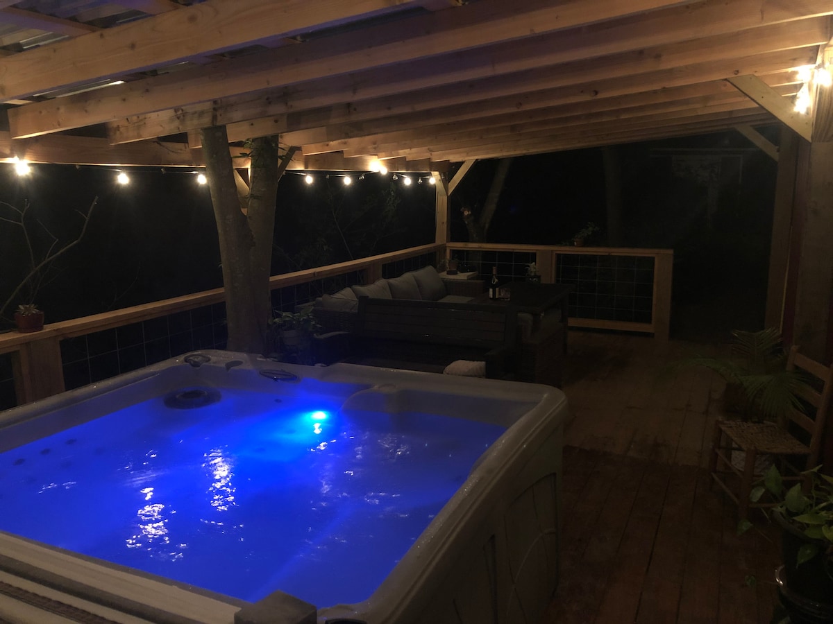 A private hot tub is illuminated in soothing blue light on a spacious wooden deck. Fairy lights hang overhead, adding a soft glow to the area. Comfortable seating is available in the corner, creating a relaxed atmosphere surrounded by nature.