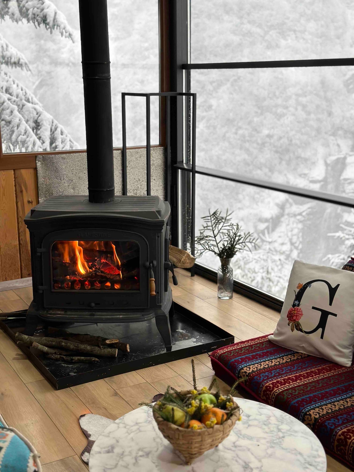 A modern wood stove provides warmth and ambiance in the living area. Nearby, a colorful patterned cushion rests against the wall, complementing a white pillow with an embroidered letter. A decorative bowl filled with fresh fruit sits on a marble table, while snow blankets the landscape outside.