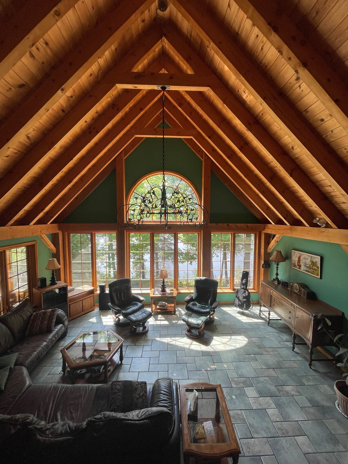 A spacious living room features high cathedral ceilings with wooden beams and a large stained glass window. The area is furnished with a dark leather couch, two armchairs, and a wooden coffee table. Natural light streams in from multiple windows, highlighting the stone floor.