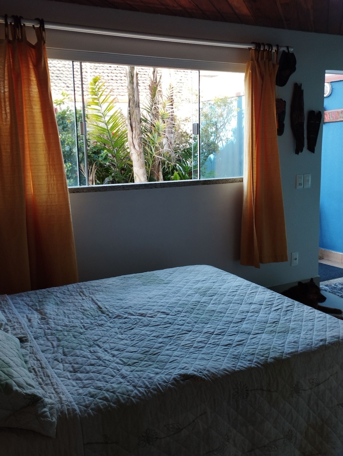 The sleeping area features a spacious bed with a light-colored quilt, positioned near a large window dressed with orange curtains. Outside, lush greenery is visible, bringing a sense of nature into the room. The bathroom is accessible nearby.