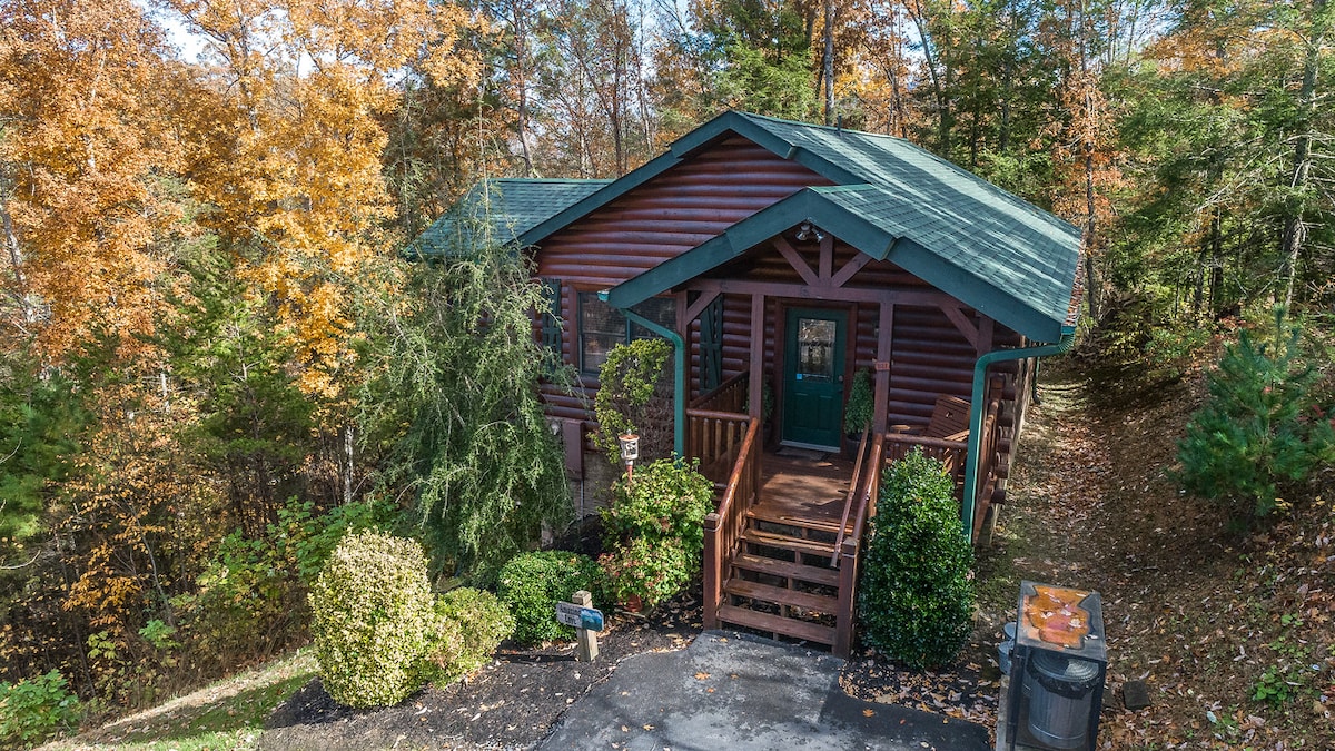 A charming cabin is nestled among vibrant trees, showcasing a rustic exterior in warm tones. The entrance features a welcoming wooden staircase and a covered porch, surrounded by neatly trimmed bushes and a gravel pathway. The sloped lot offers a natural and serene setting.